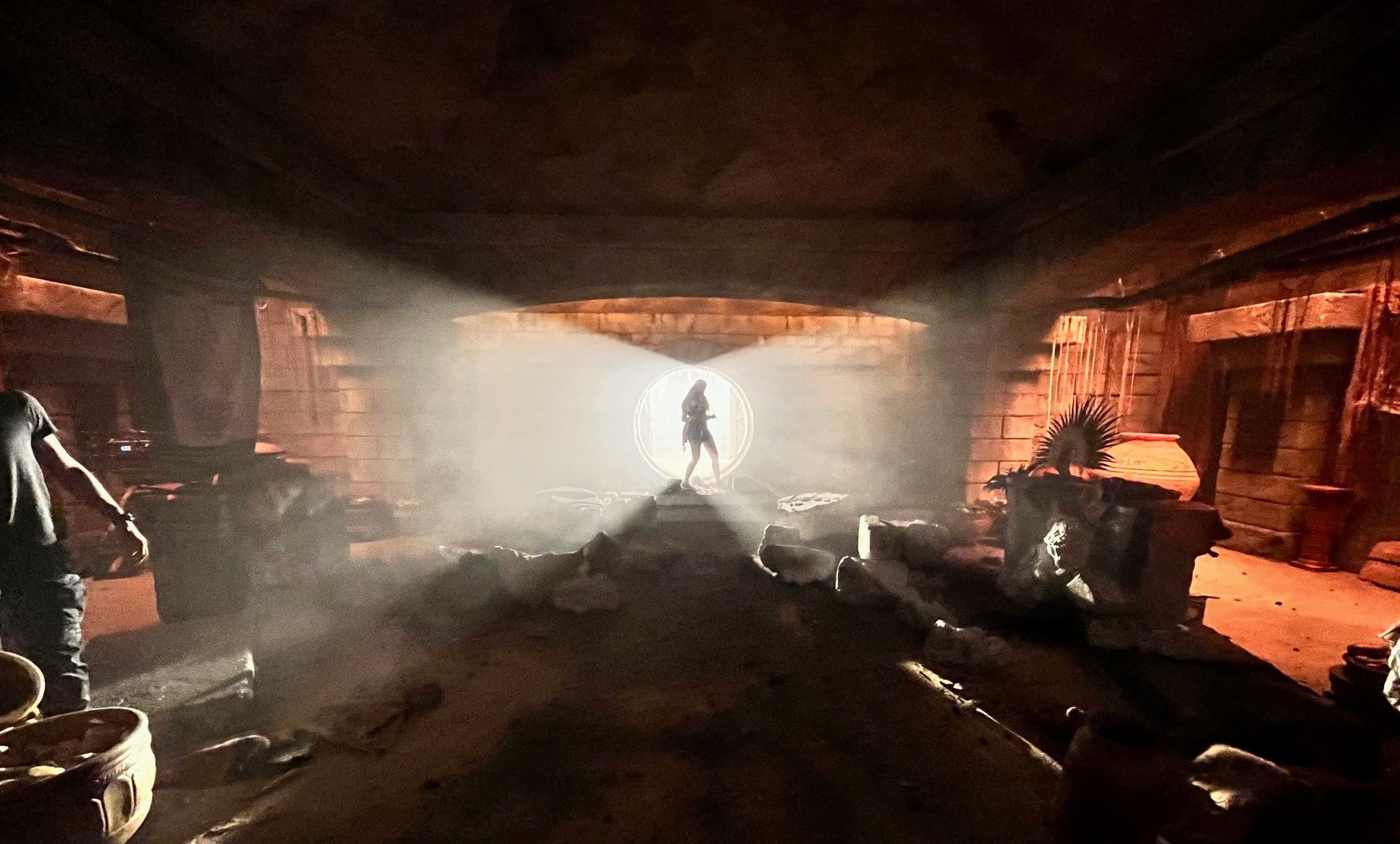 Silhouette of a woman standing in a circular doorway with a bright light shining behind, set in an indoor, cave-like environment.