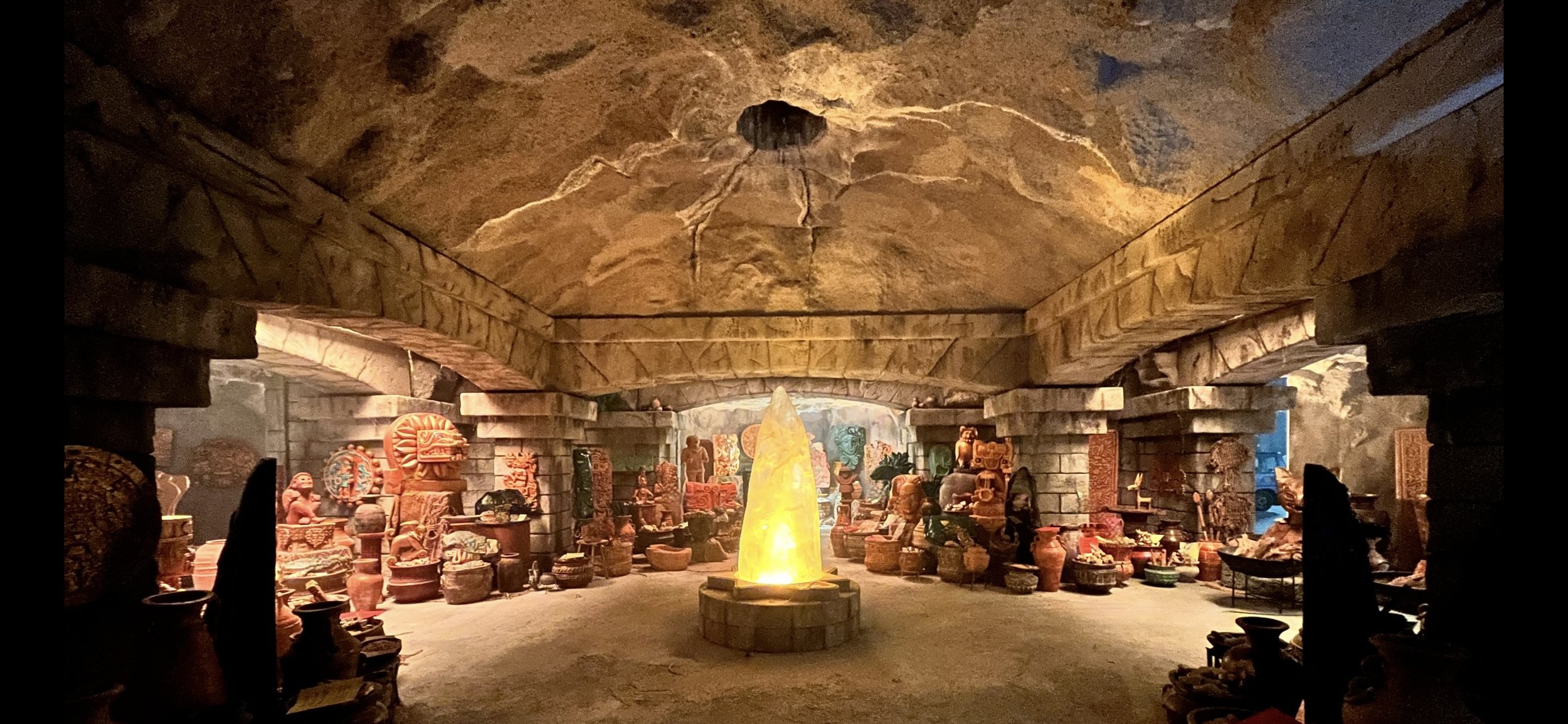 Interior shot of a room filled with cultural artifacts and pottery surrounding a glowing crystal-like structure, creating a mystical ambiance.