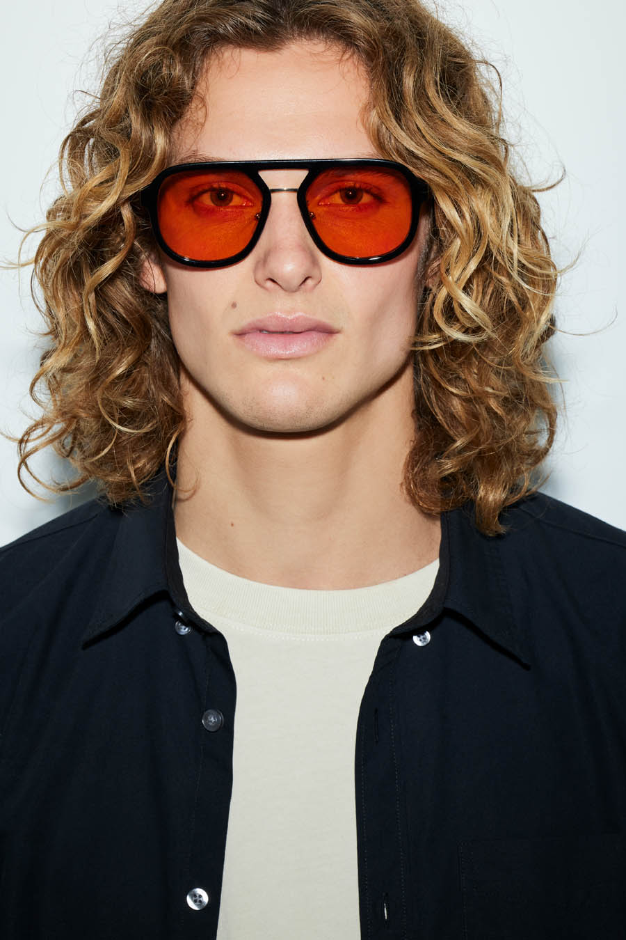 Headshot of a curly-haired man wearing bright orange sunglasses and a dark blue collared shirt against a white studio backdrop.