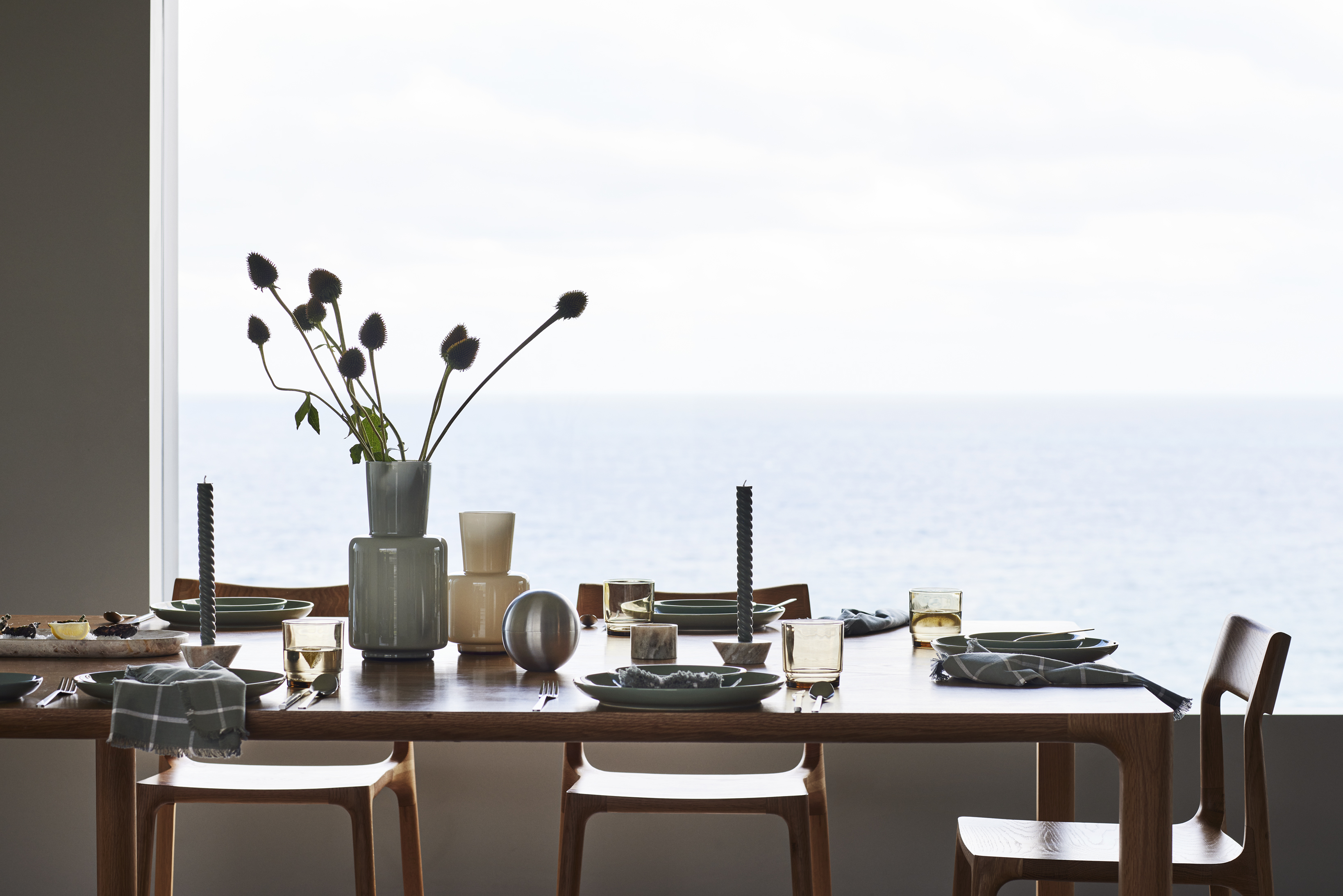 A wooden table is set with plates, glasses, silverware, and flower arrangements in a room with large window overlooking a calm ocean view.