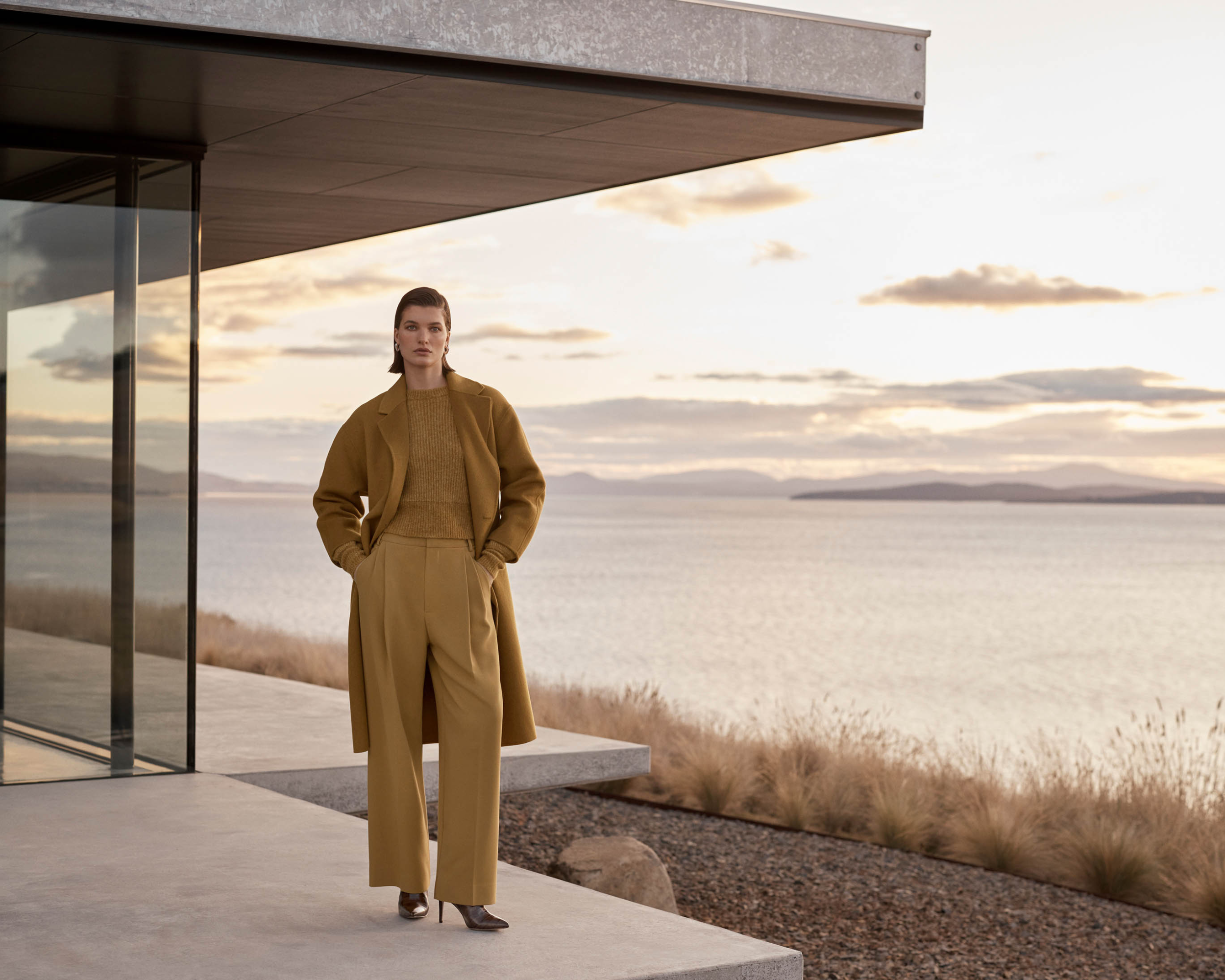 Woman poses on concrete patio wearing brown coat, sweater, and pants overlooking the ocean, showcasing fashion and lifestyle.