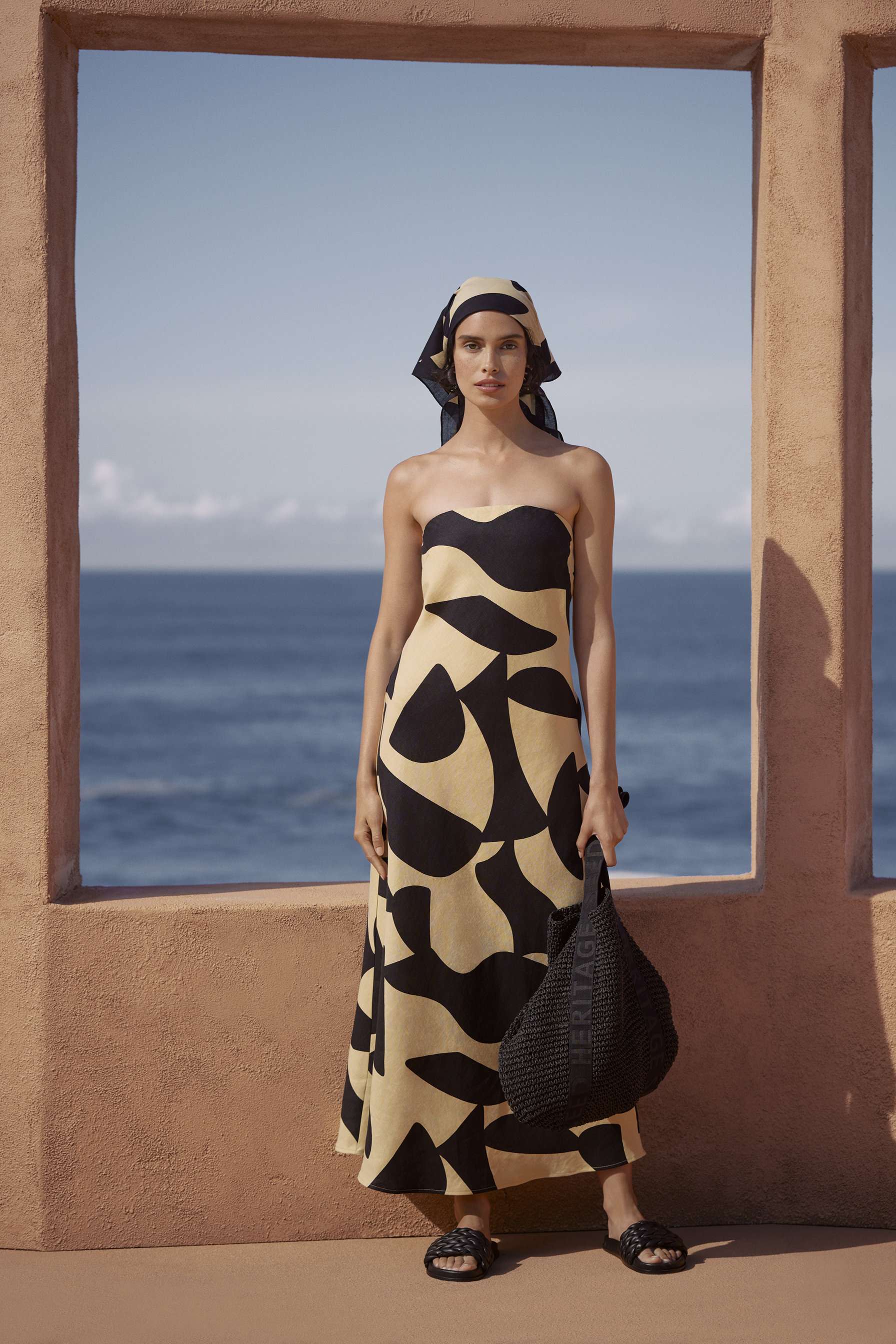 A woman in a patterned dress and headscarf stands by a window overlooking the sea, holding a black tote bag.