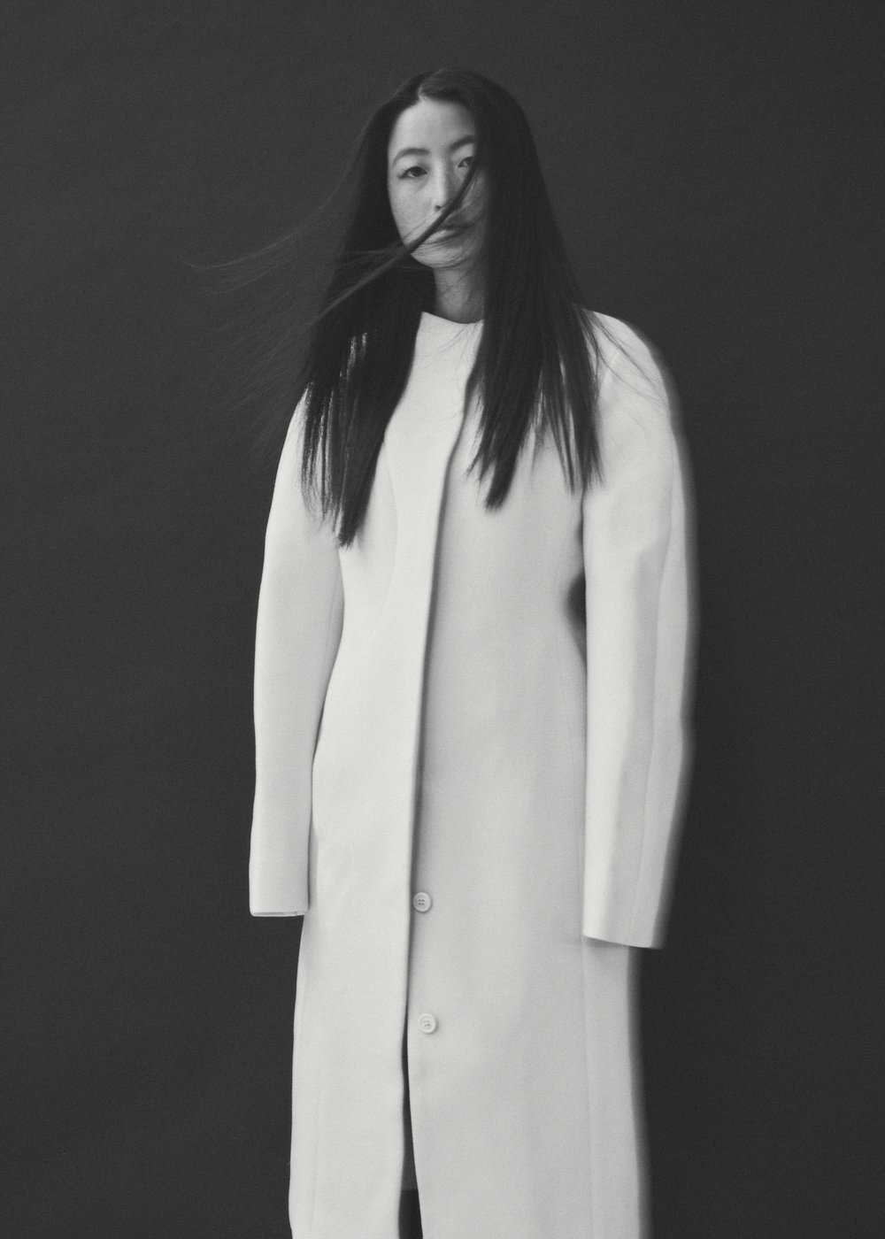 Elegant portrait of an Asian woman with long black hair wearing a white coat against a dark grey background in a studio setting.