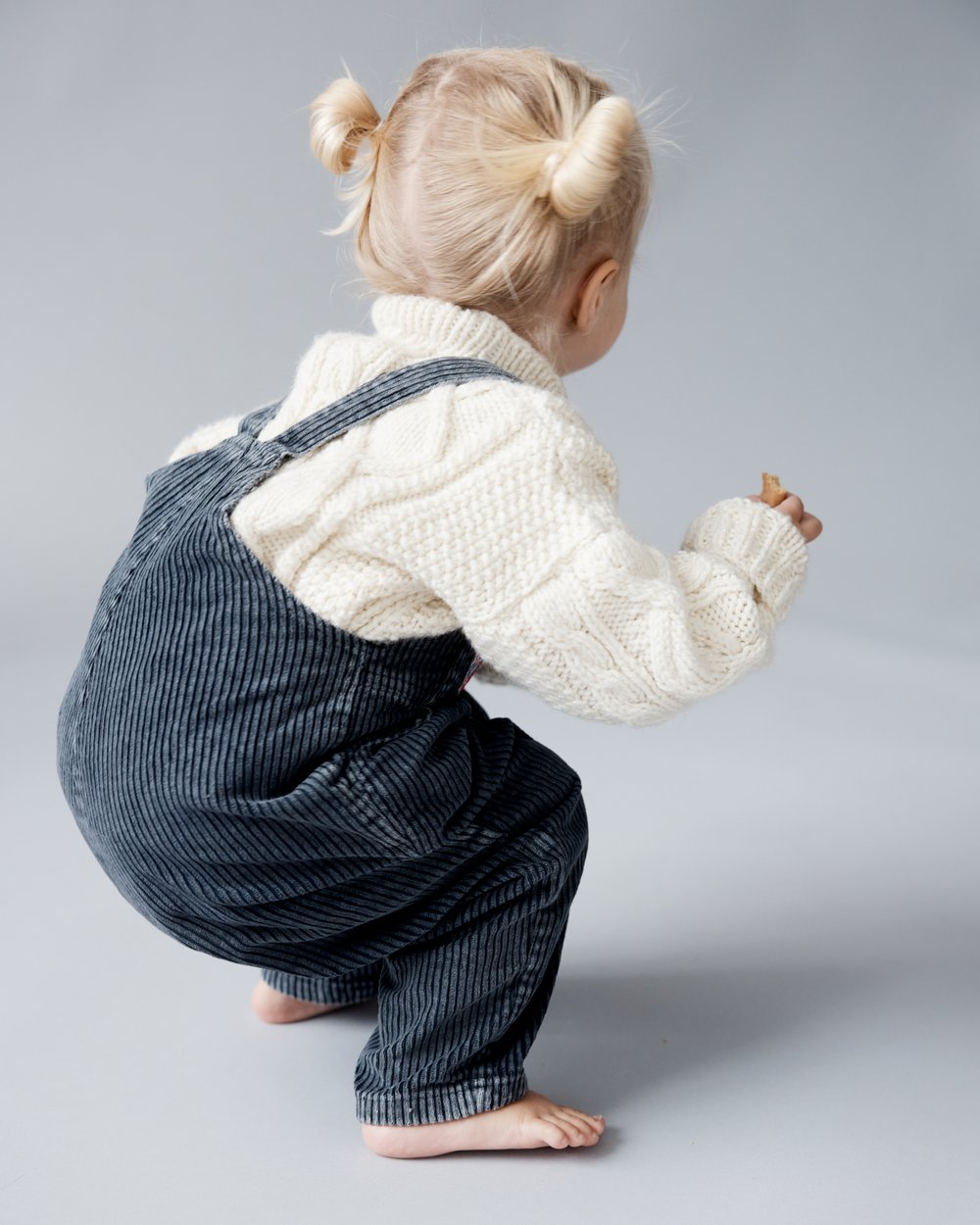 A young child with blonde hair in pigtails, wearing a sweater and overalls, is crouched with their back to the camera in a studio setting.