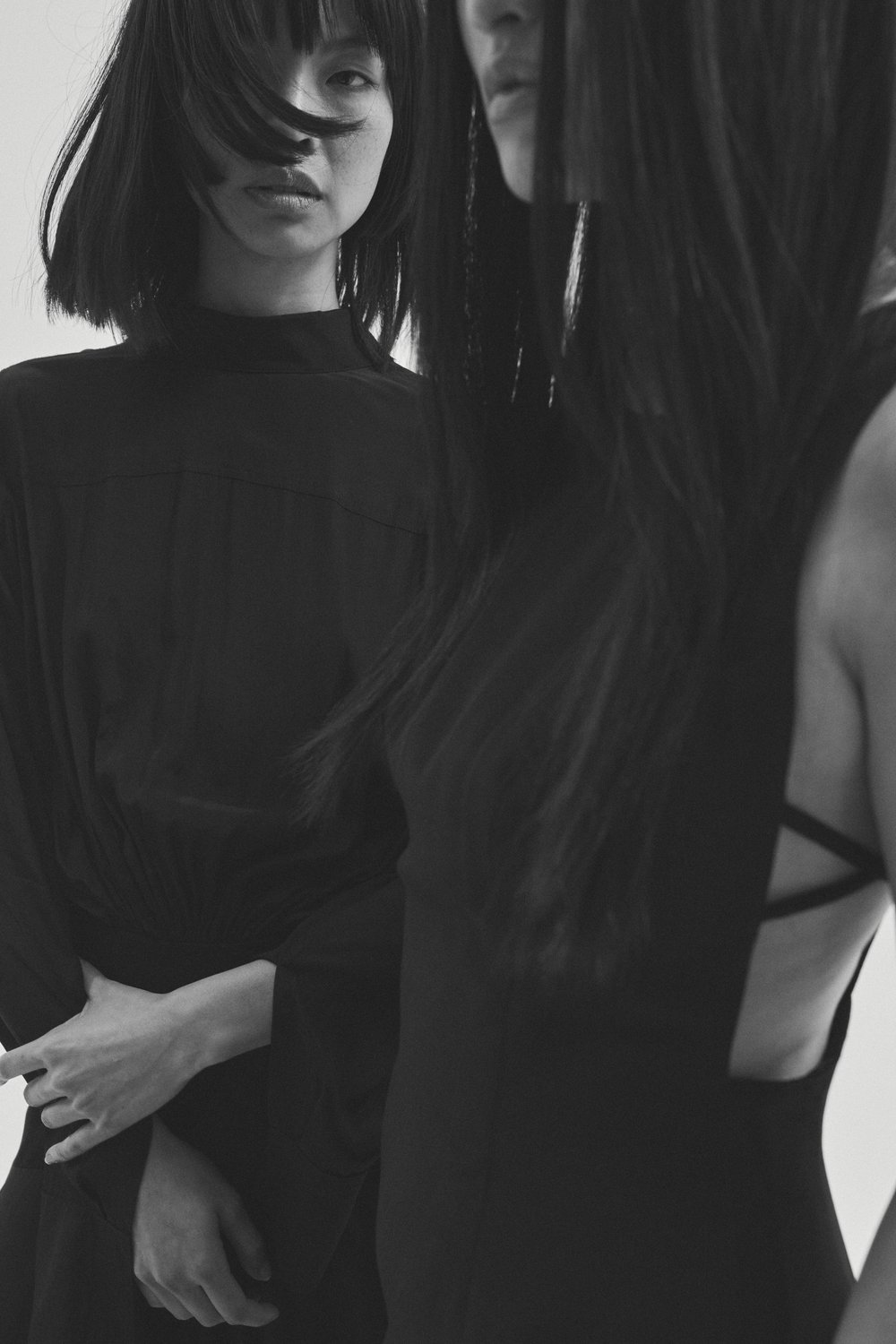 Two dark-haired women in black dresses pose in a minimalist studio setting. One gazes at the camera, the other is partially obscured.