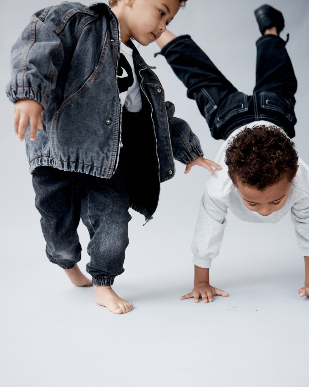 Two young children pose playfully; one does a handstand while the other stands near in a denim jacket and jeans on a light backdrop.