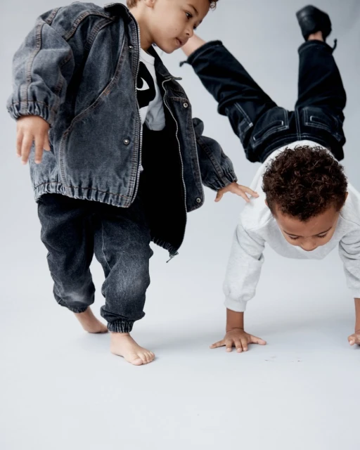 Two young children pose playfully; one does a handstand while the other stands near in a denim jacket and jeans on a light backdrop.