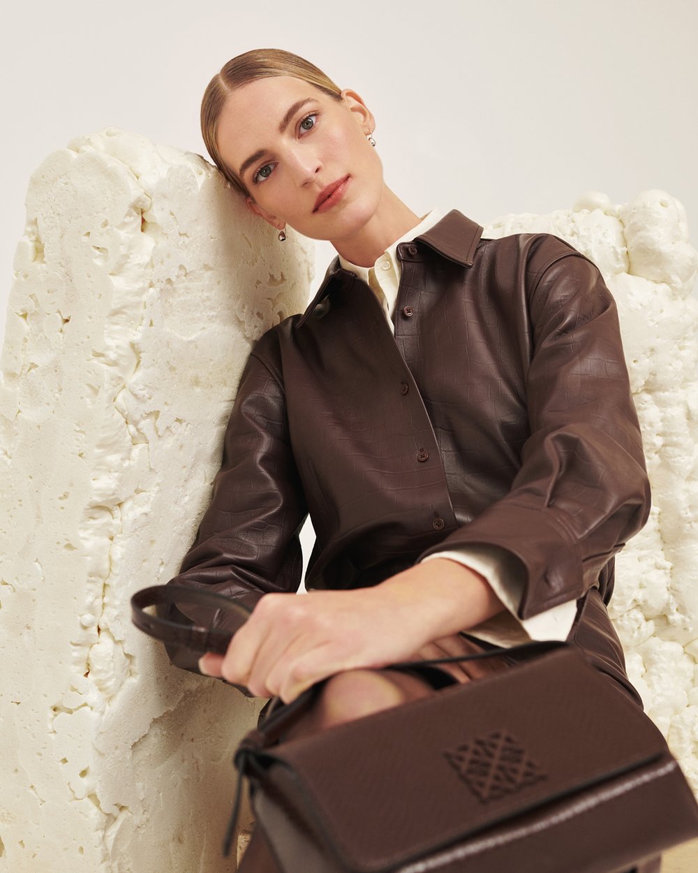 Fashionable adult female model poses with a brown leather handbag in a studio with a textured off-white background for a beauty and fashion shoot.
