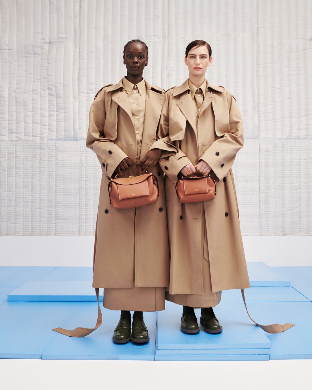 Two fashion models in tan trench coats hold matching handbags. They stand on a blue platform against a light-colored, patterned backdrop.