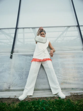 A woman in white pants and sleeveless top holds a handbag above her head in an indoor greenhouse environment.