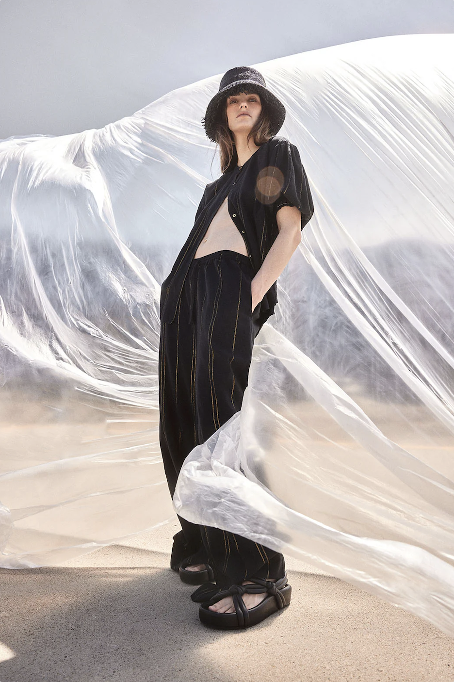 A woman wearing black, striped outfit with platform shoes poses outdoors in front of translucent fabric for a fashion editorial shoot.