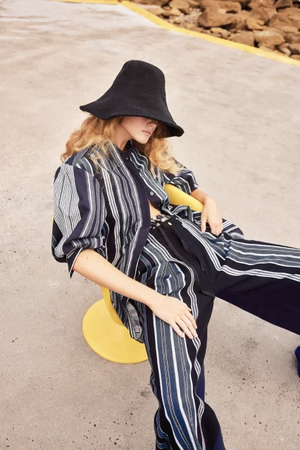 Adult woman in striped shirt and pants seated on yellow stool with rock formations in background wearing wide brim hat.