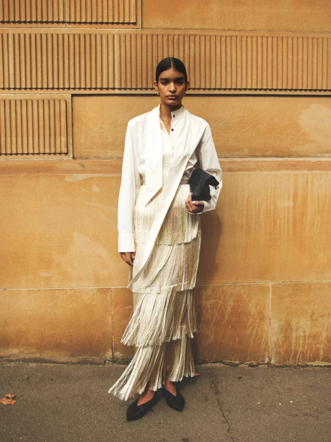 Woman in a white blouse and champagne-colored layered fringe skirt holds a black clutch while posing in front of a patterned wall.