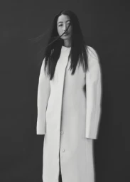 Monochrome portrait of an Asian woman with long dark hair wearing a white structured coat against a dark background.