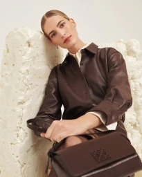 Woman in brown leather shirt leaning against a sculpted white textured backdrop holding a dark handbag
