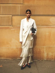 Model standing against a textured stone wall wearing a white collared shirt and layered fringe skirt holding a dark clutch.