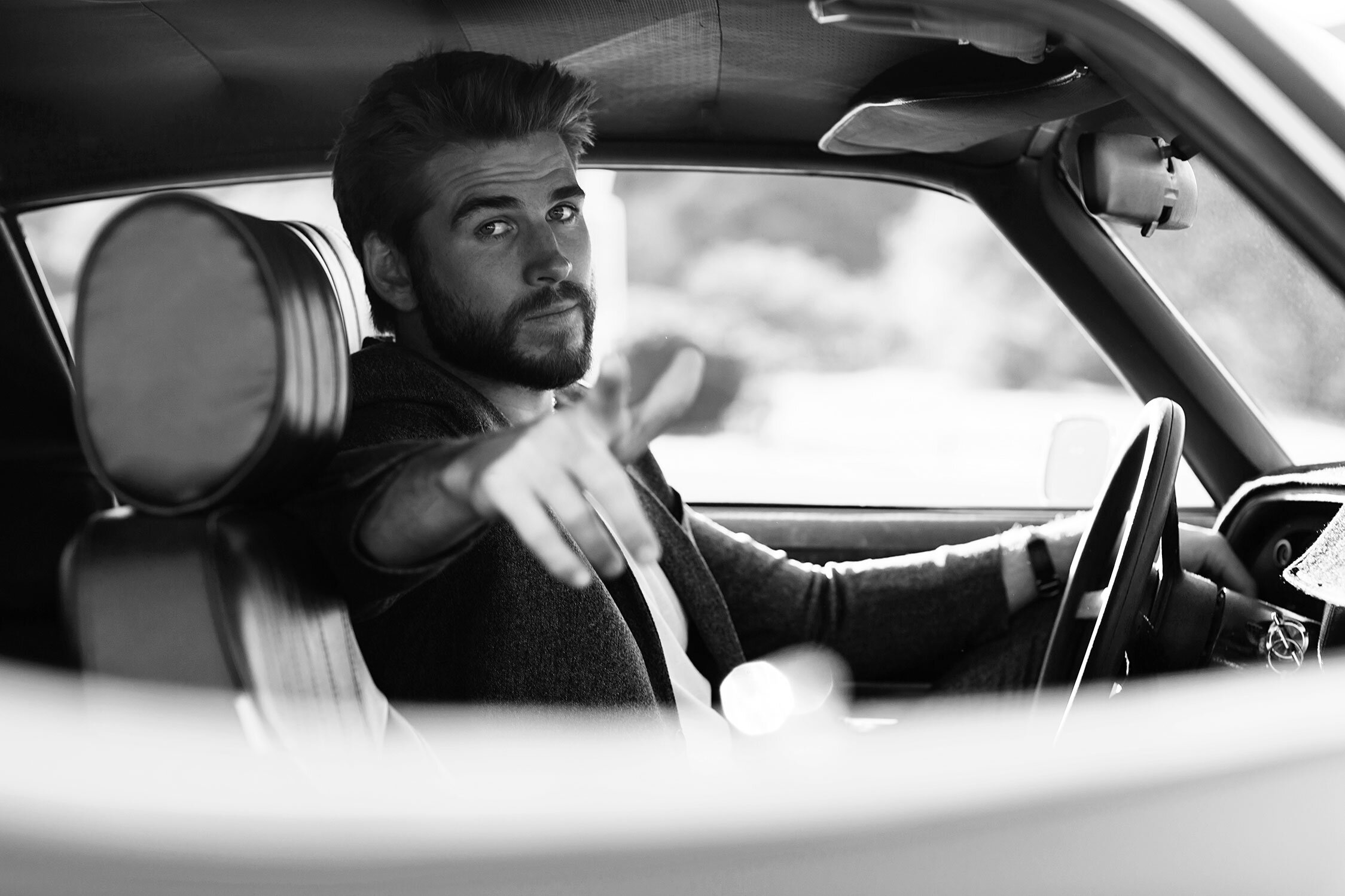 Black and white portrait of an adult male inside a car pointing toward the camera with a shallow depth of field and outdoor setting.
