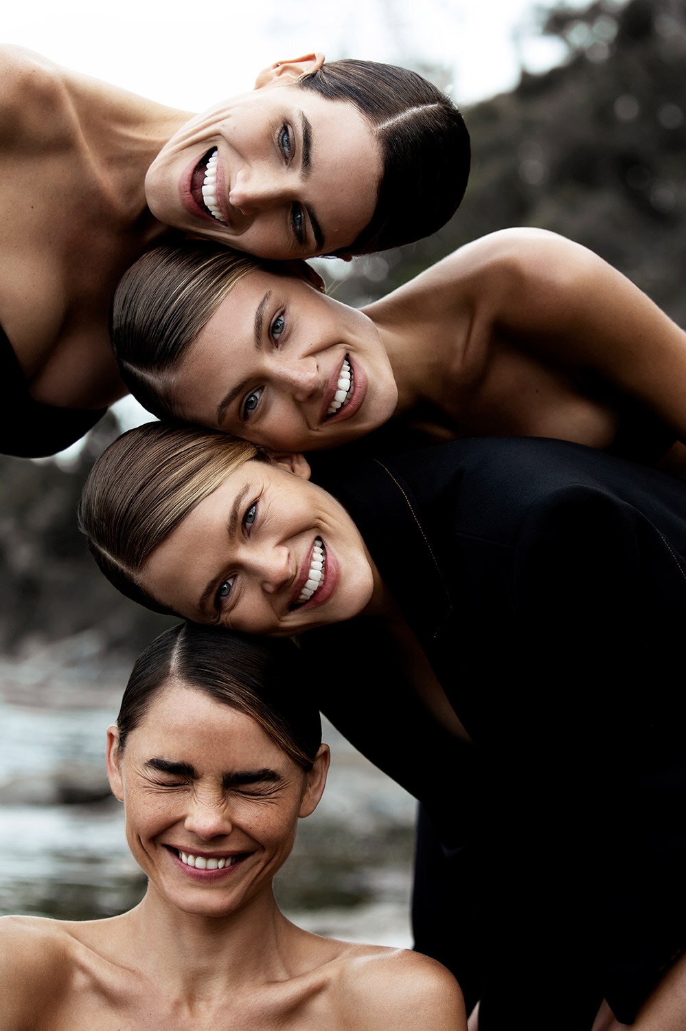 Four smiling women pose close together in an outdoor setting, creating a tower of heads with varying skin tones and dark hair.