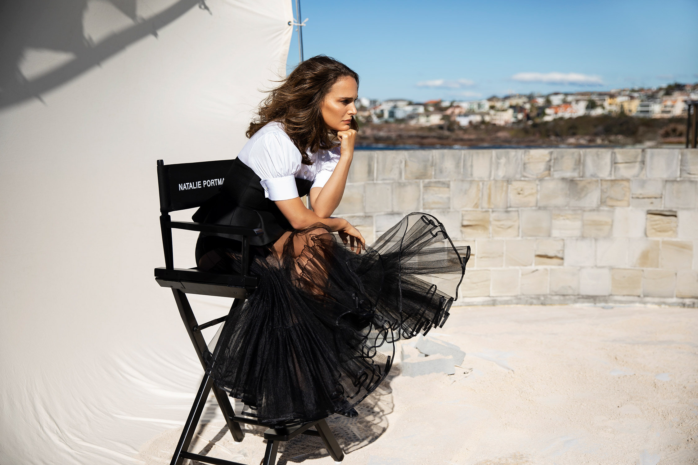 Natalie Portman poses in a black tulle dress with a white top while sitting on a director's chair outdoors, with a coastal cityscape backdrop.