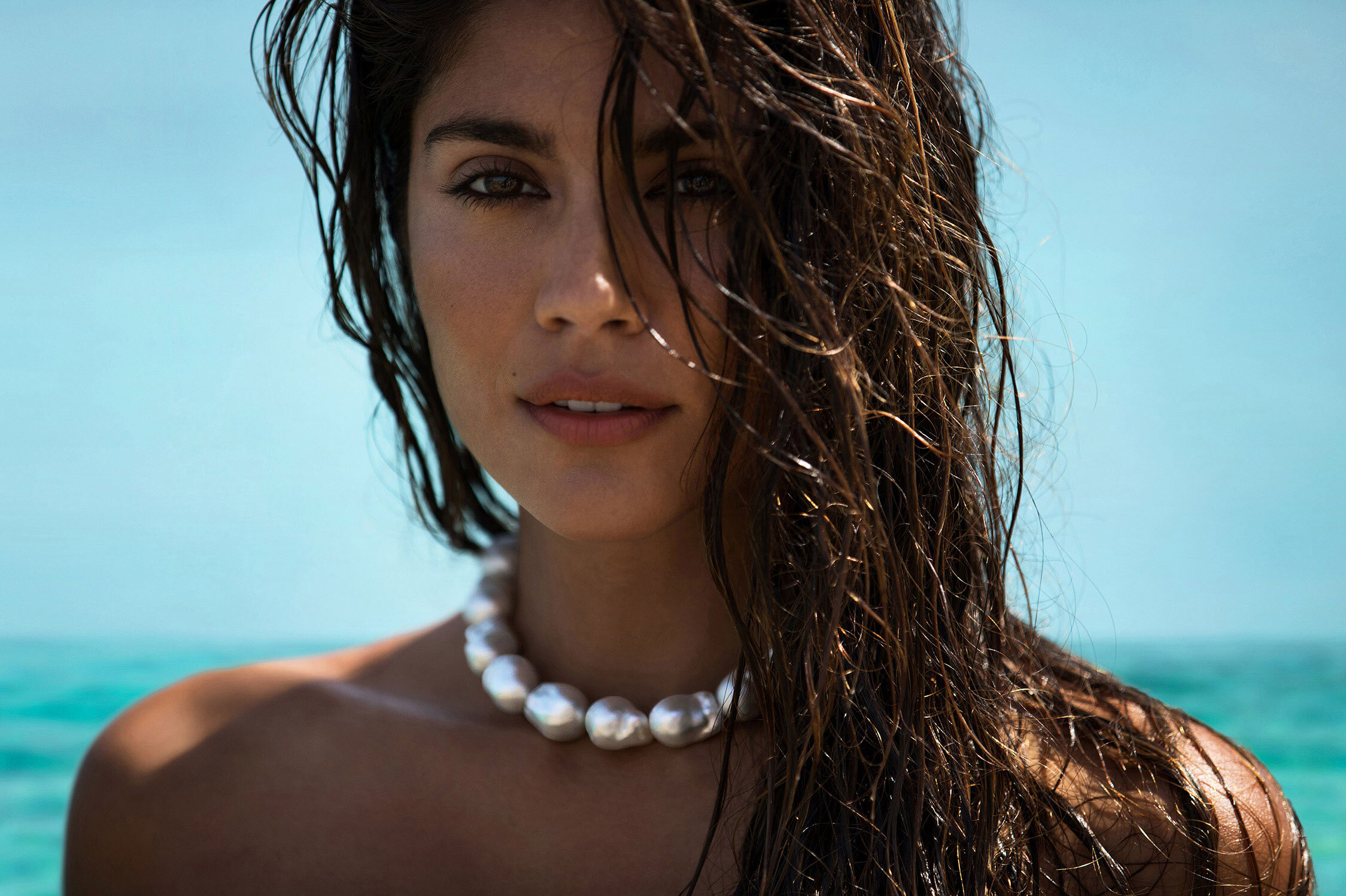 Close-up of a brunette woman with wet hair and pearl necklace against a turquoise water background on a sunny day.