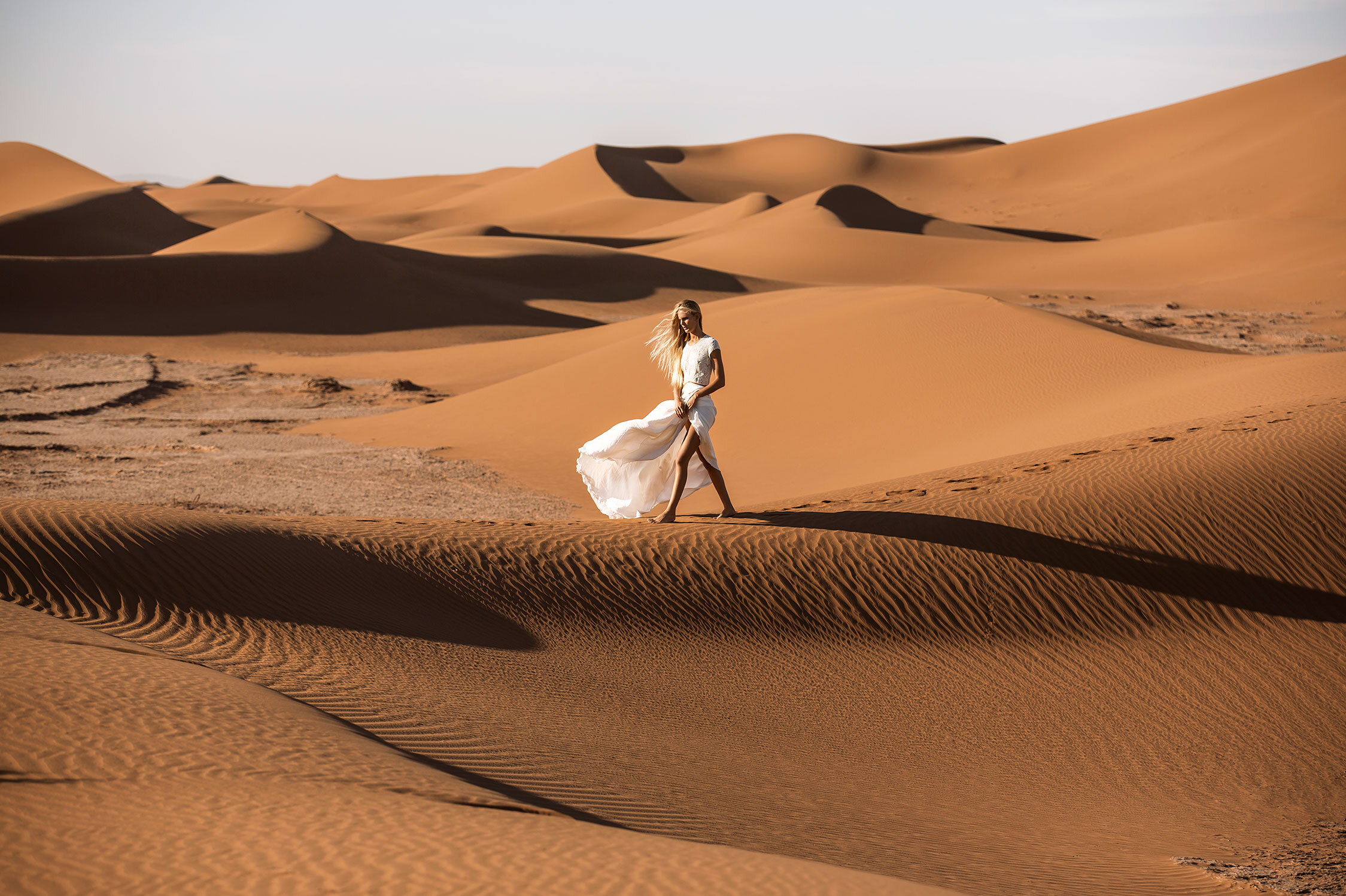A woman in a flowing white dress confidently walks on a sand dune in a vast desert landscape with textured sand and warm colors.