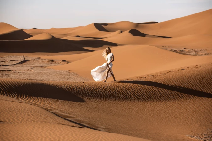 Woman in a long white dress walking across expansive sand dunes under bright sunlight