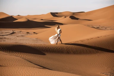 Woman in a long white dress walking across expansive sand dunes under bright sunlight