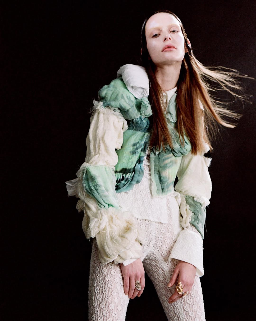 A fair-skinned woman with long brown hair poses in a studio wearing a light-colored top, jacket, and pants with rings on her fingers.