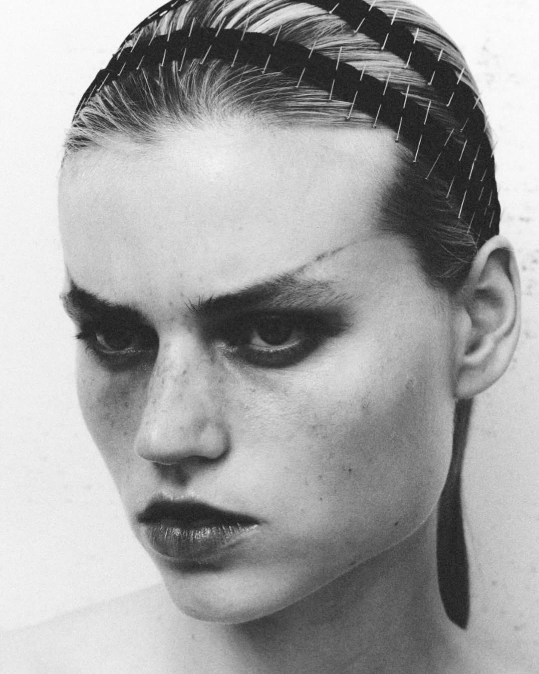Striking black and white portrait of a young adult female model with dark eye makeup, slicked-back hair, and a unique headpiece in a studio setting.