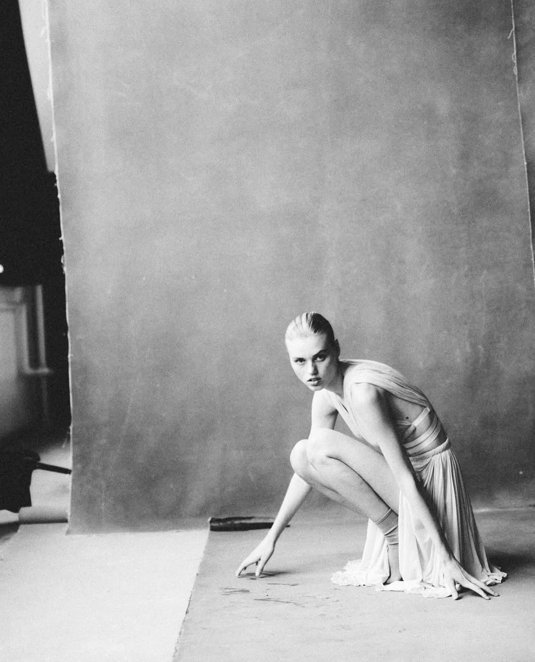 Black-and-white studio shot of a fair-skinned adult woman, crouching, wearing a dress with a grey backdrop, evoking a high-fashion editorial style.