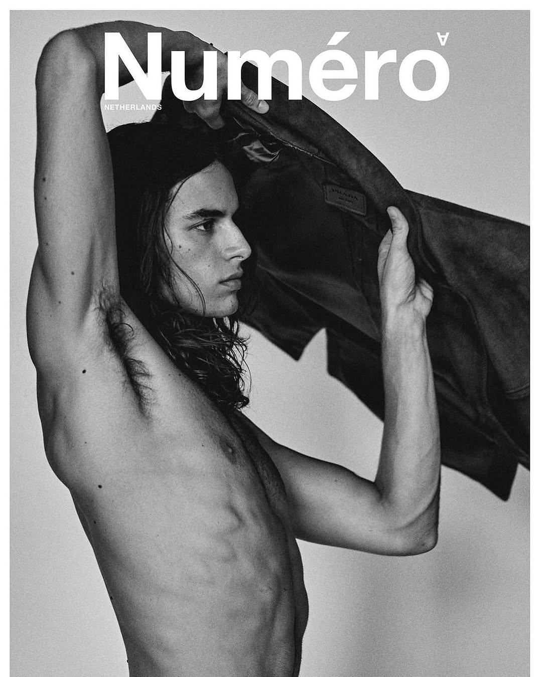 Monochrome fashion shot of a shirtless male model holding a jacket with text overlay of 'Numéro'.