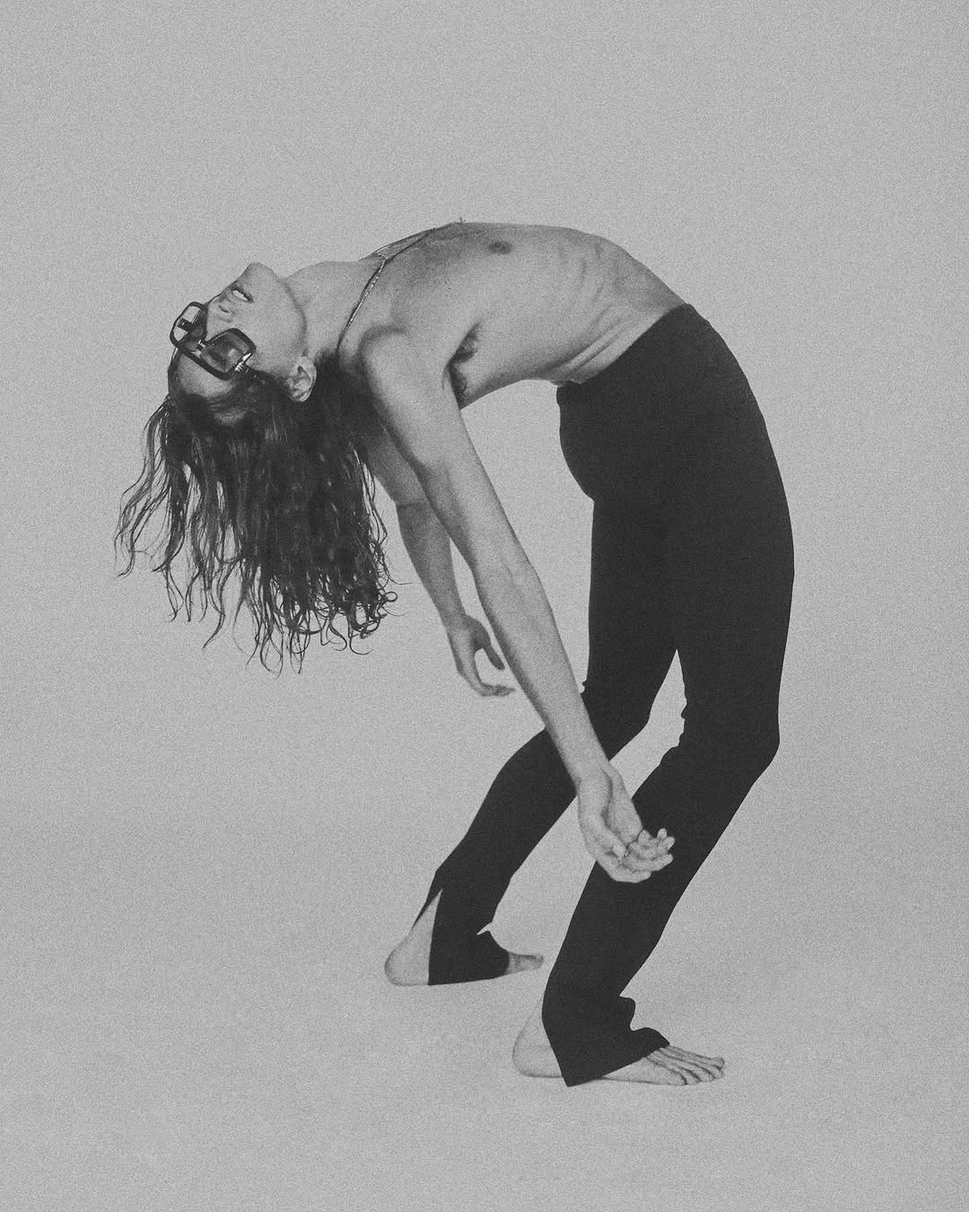 Monochromatic portrait of a long-haired person in split-hem pants, arched backwards while wearing dark glasses against a plain background.