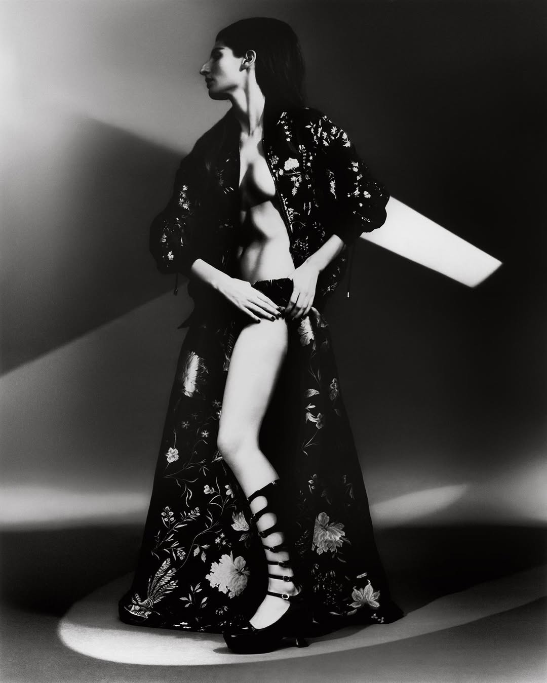 Black-and-white studio shot of a woman in a floral kimono-style robe, underwear, and gladiator sandals, with dramatic lighting and a posed profile.
