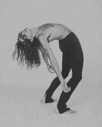 Monochrome fashion portrait of a long-haired model arching backward wearing dark sunglasses and slim black trousers.