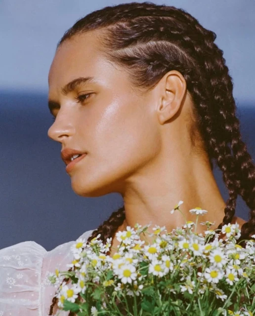 A woman with braided hair holds a bouquet of white daisies against a blue background in a close-up, eye-level, outdoor fashion portrait.