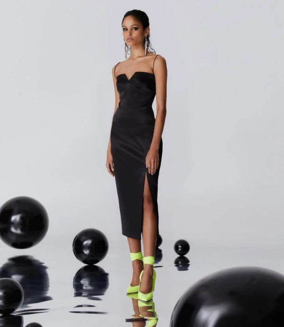 Elegant woman in a black midi dress with a thigh-high slit and bright yellow heels poses among black spheres in a photo studio.