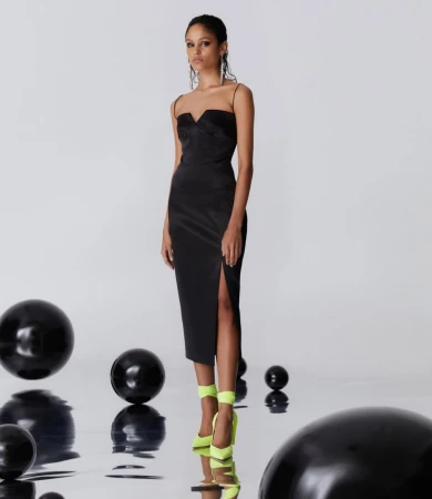 Elegant woman in a black midi dress with a thigh-high slit and bright yellow heels poses among black spheres in a photo studio.