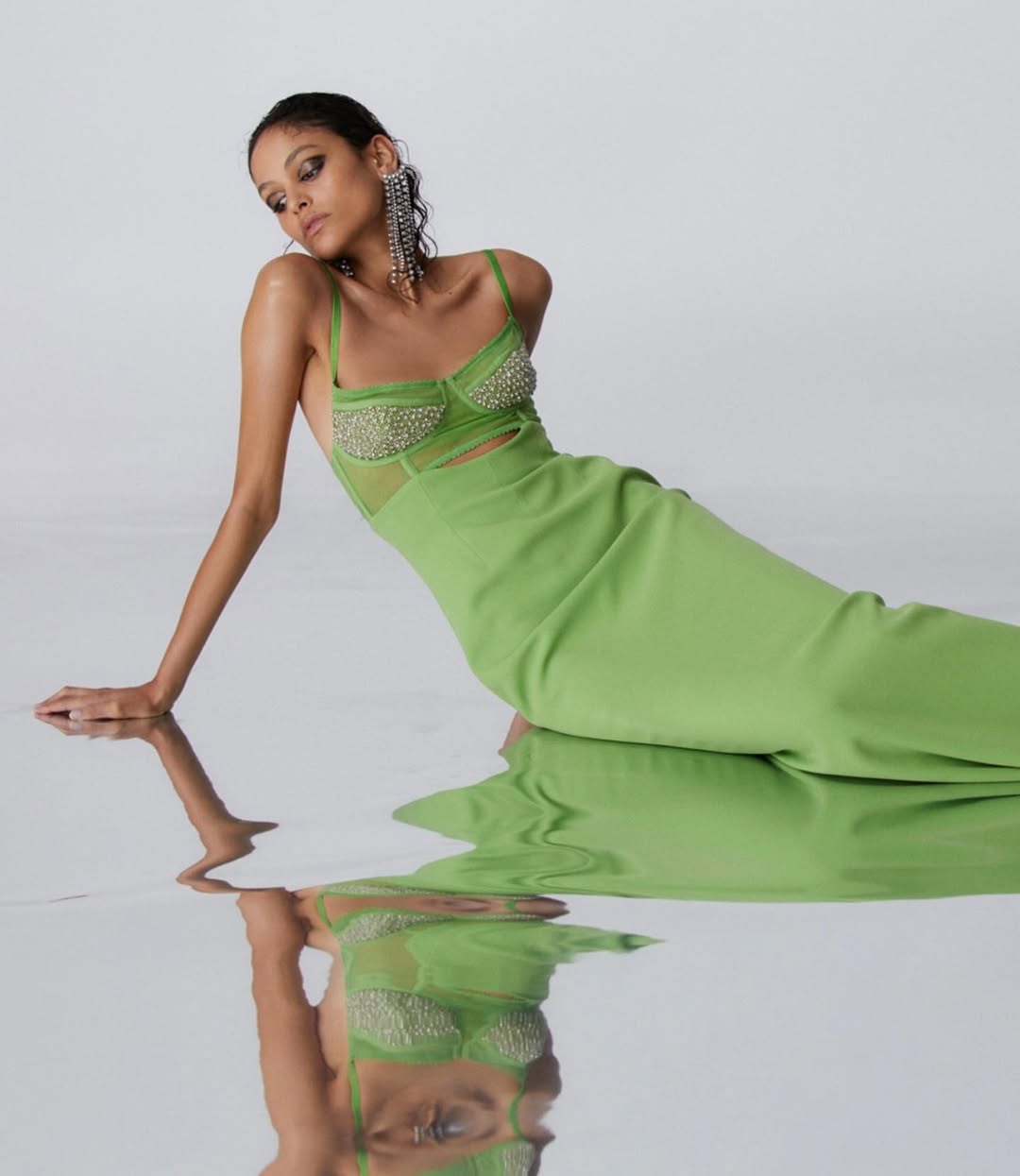 Woman in light green dress reclines against mirrored surface in studio. She is wearing large dangling earrings and her reflection is visible.