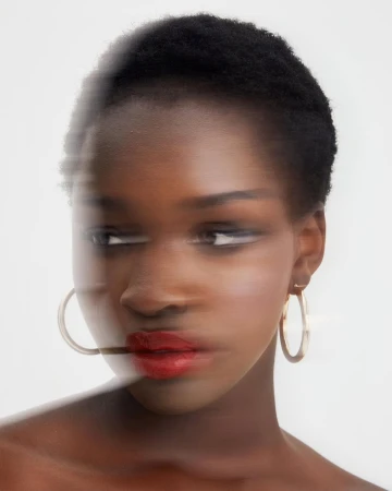 Close-up portrait of a Black woman with short hair wearing gold hoop earrings and red lipstick against a white background