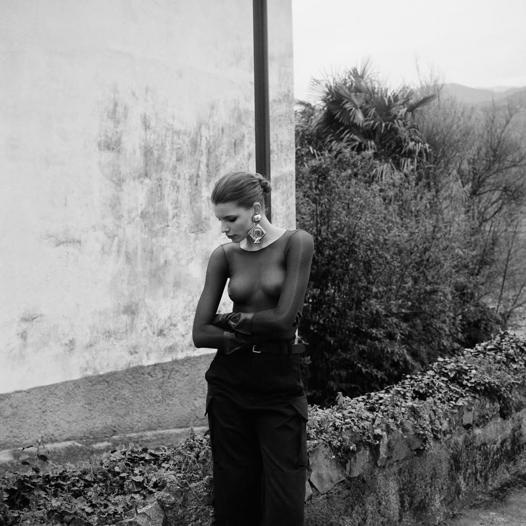 Black and white portrait of a young woman with bare midriff wearing large earrings, dark top and pants in outdoor setting.