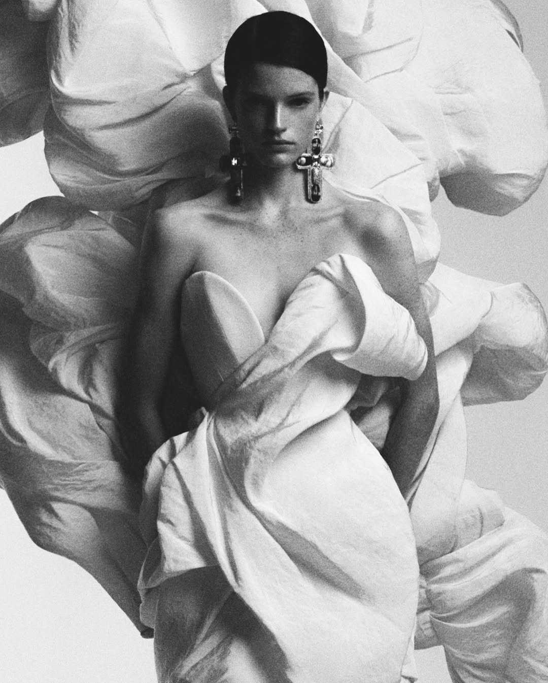 Black and white fashion portrait of a woman with earrings wrapped in billowing white fabric against a soft gradient background.