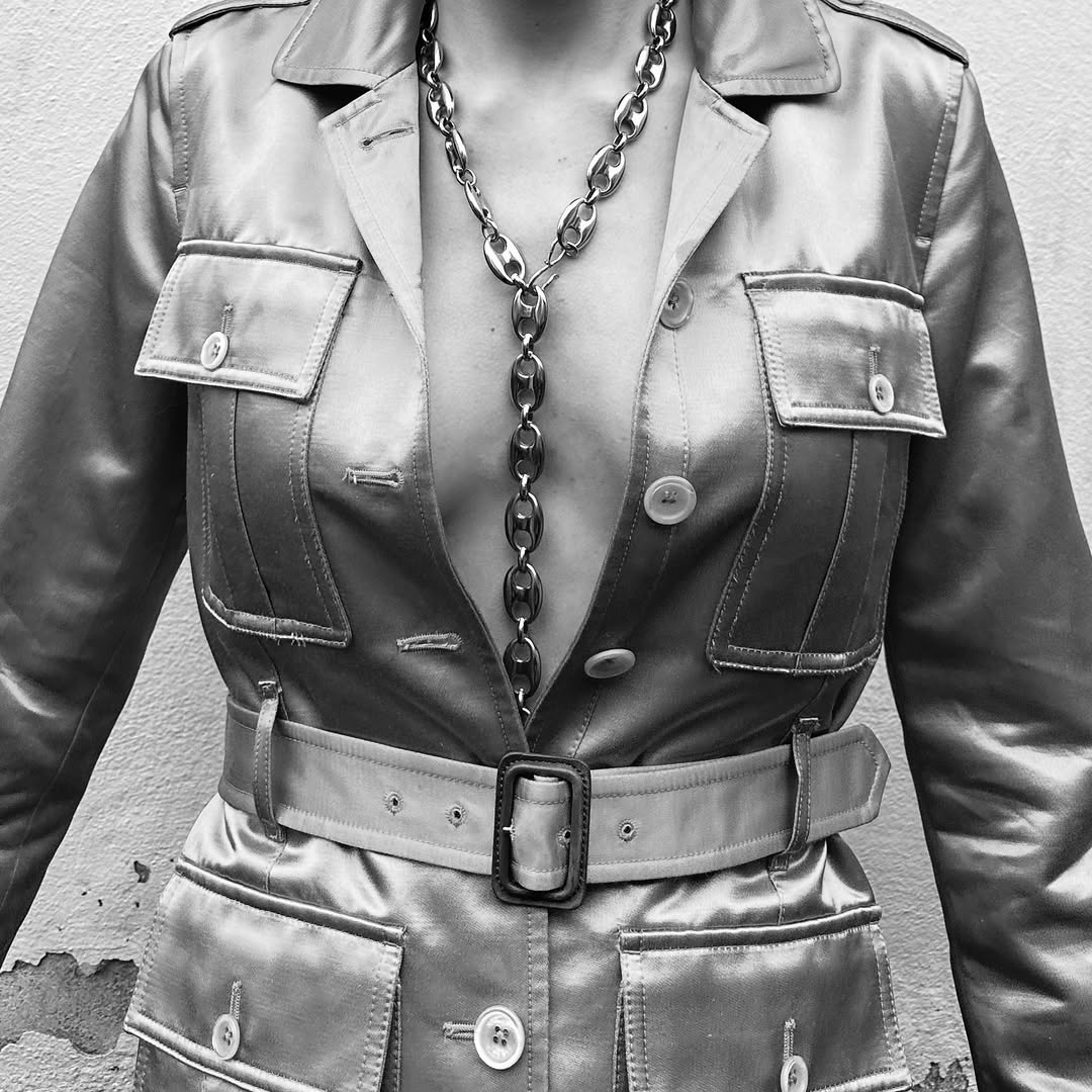 A silver chain necklace worn with a shiny gold jacket and matching belt, close-up shot against a neutral textured background.