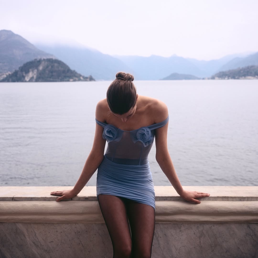 Woman in a blue dress poses on a marble balcony with a mountainous lake background, creating an elegant fashion shot with a scenic view.