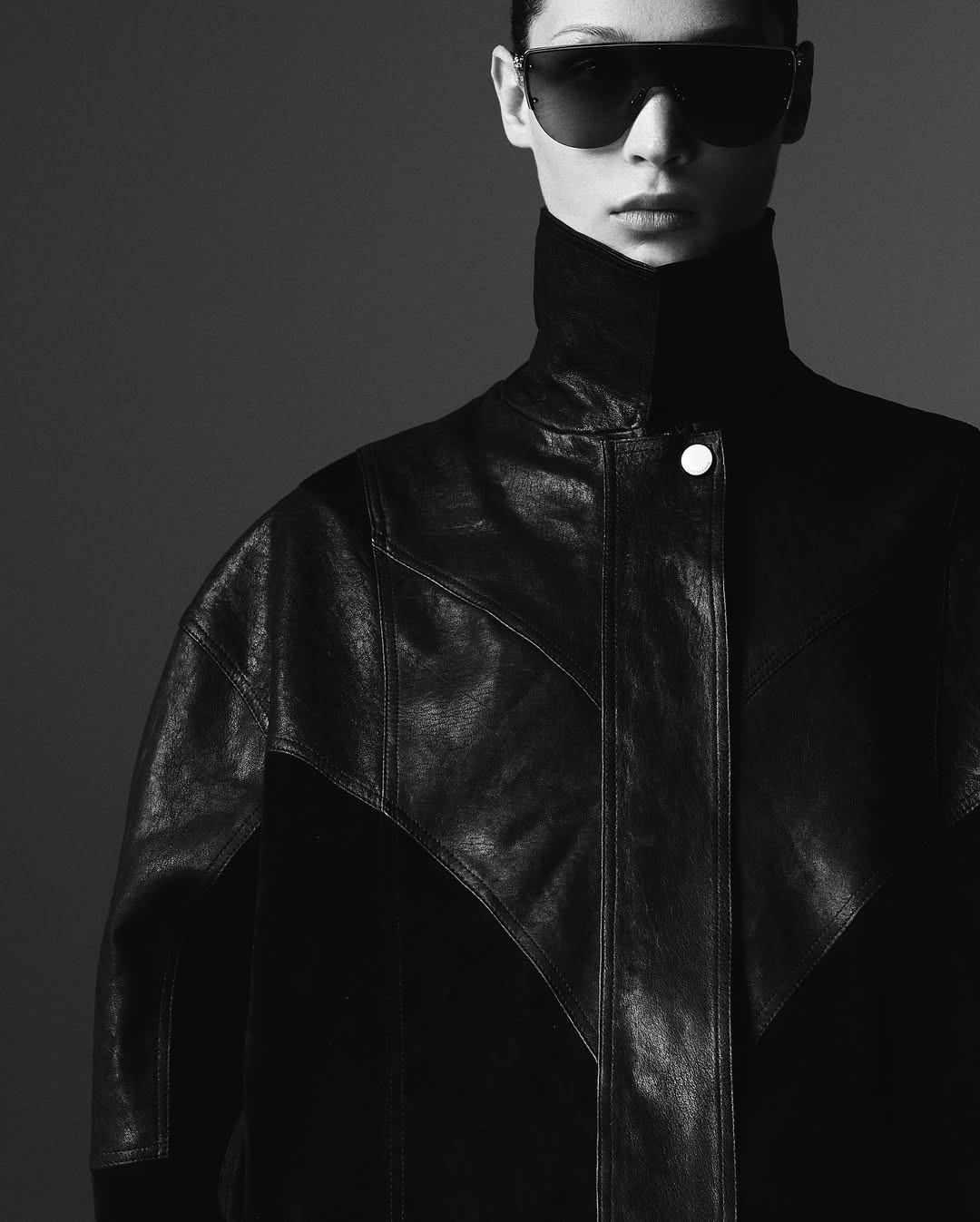 Monochrome portrait of a fashionably dressed woman wearing oversized sunglasses and a black leather jacket against a grey backdrop.