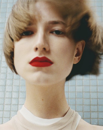 Close-up portrait of a fair-skinned young woman with red lipstick, short brown hair and a neutral expression set against a tiled background.