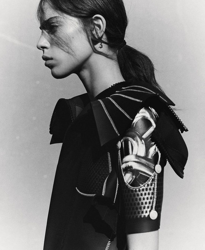 Monochrome portrait of a woman with dark hair in a ponytail, wearing a detailed black outfit with intricate shoulder embellishments.