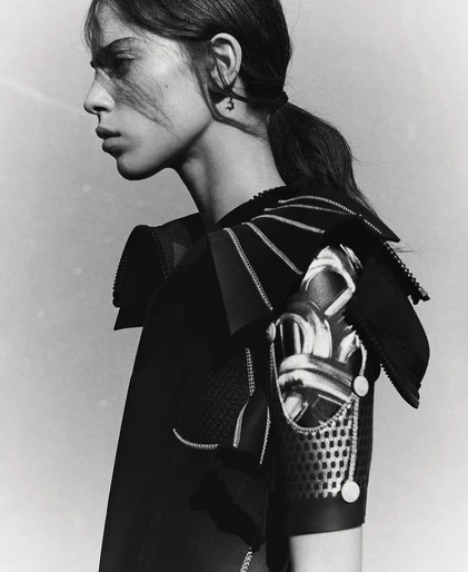 Monochrome portrait of a woman with dark hair in a ponytail, wearing a detailed black outfit with intricate shoulder embellishments.
