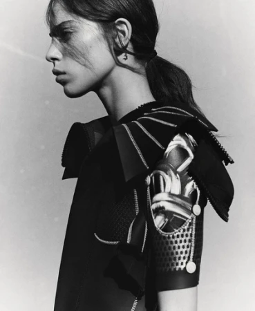 Monochrome portrait of a woman with dark hair in a ponytail, wearing a detailed black outfit with intricate shoulder embellishments.