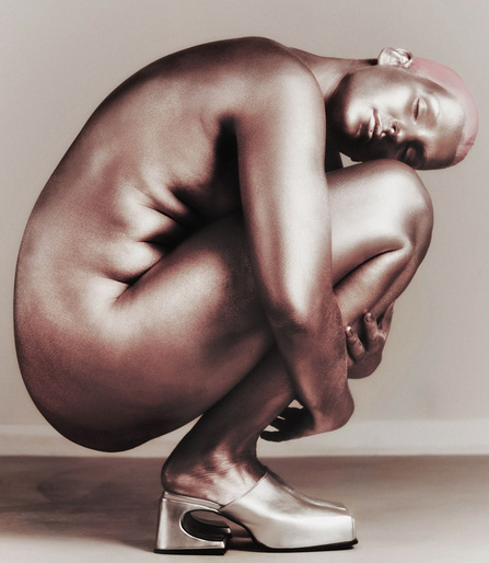 Nude adult female with metallic skin crouches with arms around legs, wearing silver shoes, in a studio shot on a beige background.