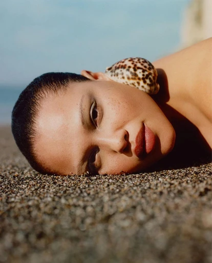 Woman with a shell on her head lies on the beach; portrait with ocean and sky backdrop. Beauty and nature combine in sunlit portrait.